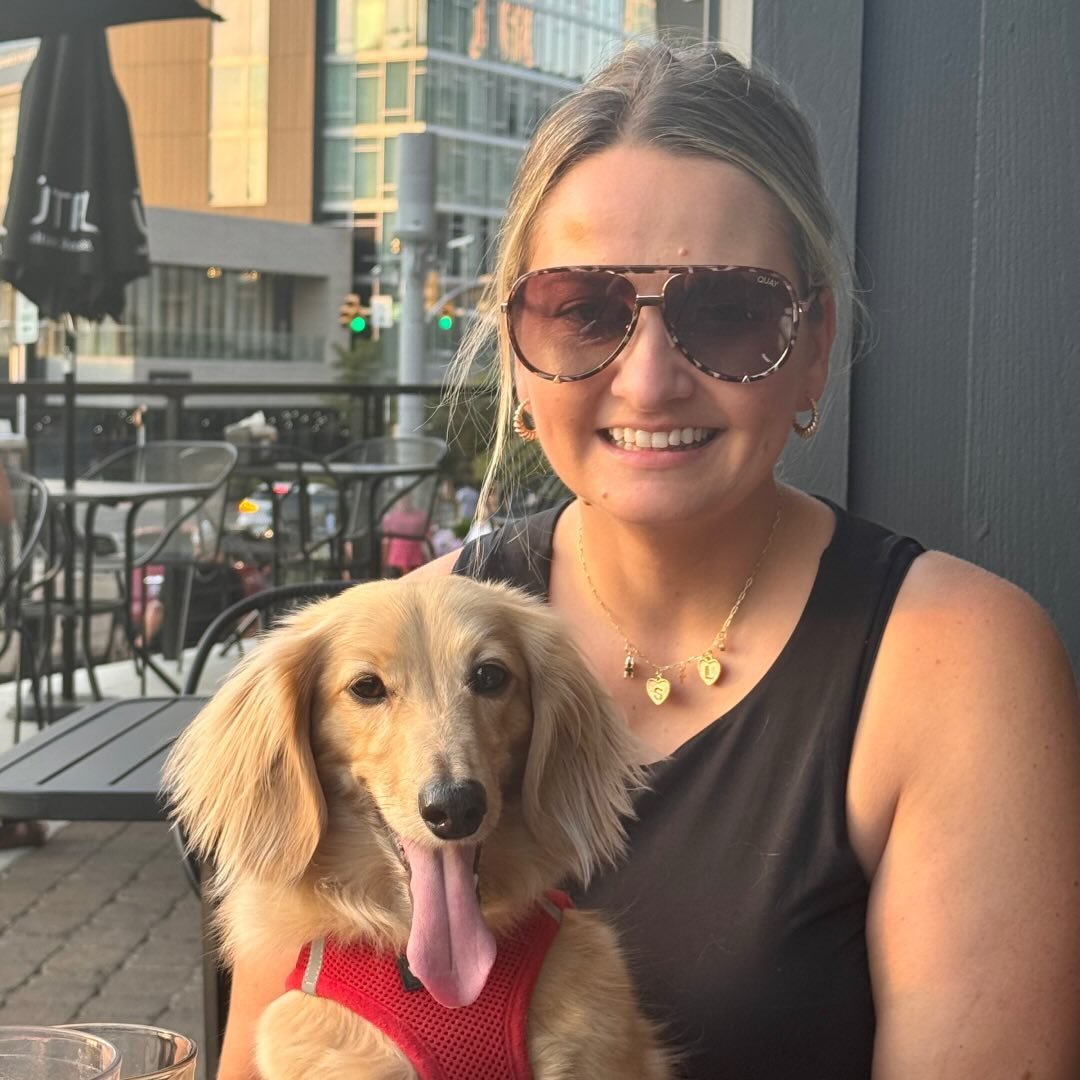 A smiling woman wearing sunglasses and a black top sits at an outdoor cafe table with a long-haired dog in her lap, dressed in a red harness. Urban buildings are visible in the background.