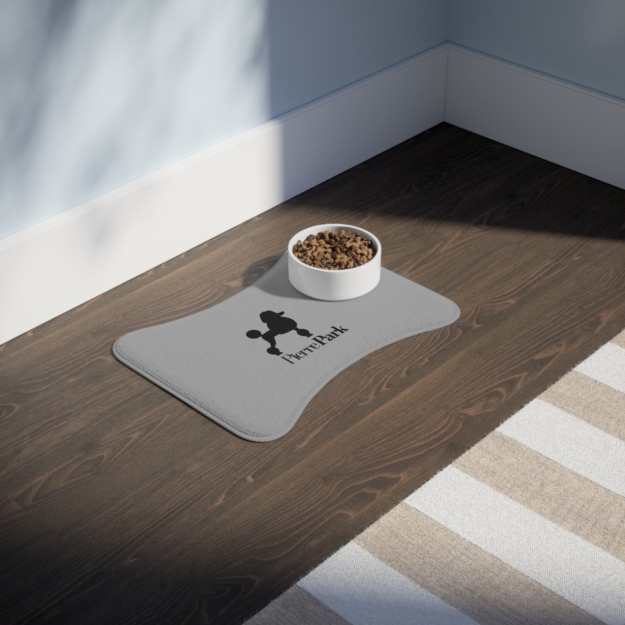 A white pet bowl filled with kibble sits on a PierrePark Dog Food Mat in Light Grey with a non-slip bottom on the dark wooden floor. The mat features a dog silhouette and Pawshack. Sunlight creates shadows, bordered by a blue and white wall.