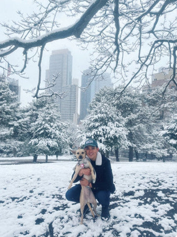 Person crouching in a snowy park, holding a dog. Snow-covered trees and buildings are visible in the background. The person is wearing a cap and winter clothing.