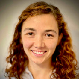 Smiling person with curly, light brown hair against a neutral background. They are wearing a light-colored top.