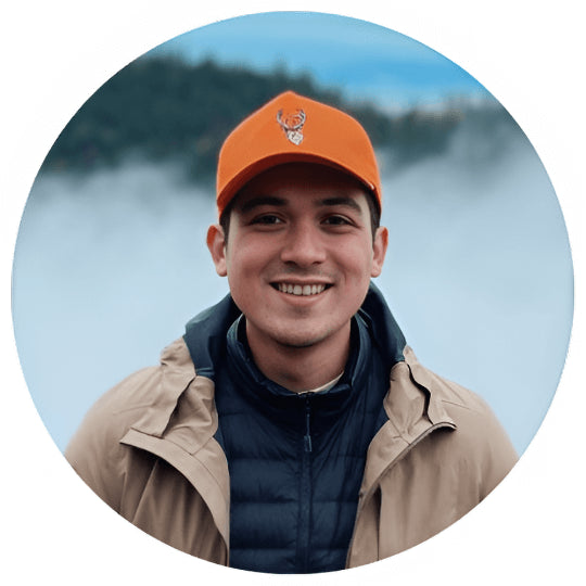 A person wearing an orange cap, beige jacket, and dark zip-up shirt smiles at the camera. The background features a fog-covered landscape with hints of greenery.