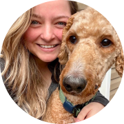 Woman smiling and hugging a light brown poodle. The poodle is wearing a blue collar, and both are looking at the camera. The background shows part of a building exterior.