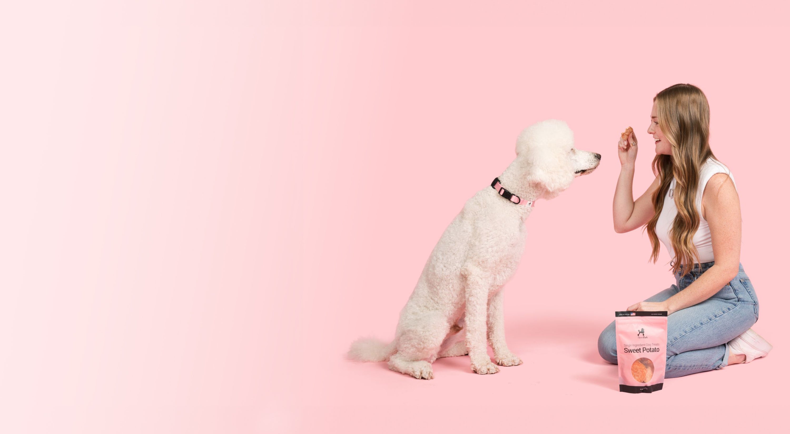 A woman with long hair, wearing a white top and jeans, is kneeling on a pink background while holding a treat in front of a large white poodle. A bag labeled Sweet Potato sits in front of them.
