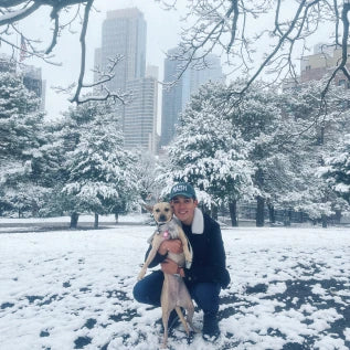 A person wearing a coat and a cap crouches and holds a dog in a snowy park. Snow-covered trees and tall buildings are visible in the background. The scene suggests a city environment in winter.