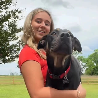 Woman in a red shirt smiling while holding a large black dog with a red collar. They are outdoors, with a grassy field and trees in the background under a cloudy sky.