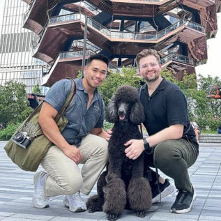 Two smiling individuals kneel beside a large, fluffy black poodle. They are in an outdoor urban area with a modern, geometric building in the background. Both are casually dressed, one wearing a camera shoulder bag.