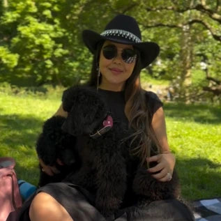 A woman wearing a black hat and sunglasses is sitting on grass in a park with two black poodles. She is smiling and dressed in dark clothing, enjoying a sunny day outside.