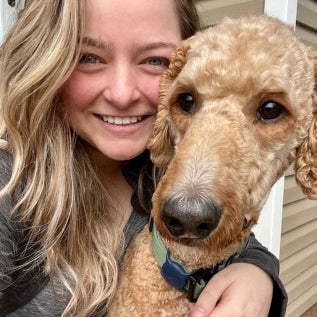 A woman with long blonde hair smiling and hugging a curly-haired dog. The dog has a reddish-brown coat and is wearing a collar. They are posed outside in front of a beige wall.