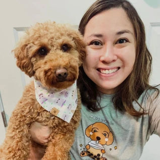 A person smiling while holding a fluffy brown dog. The dog is wearing a white bandana with a pattern. The person is wearing a gray shirt featuring a cartoon image of a similar-looking dog. They are standing in front of a white door.