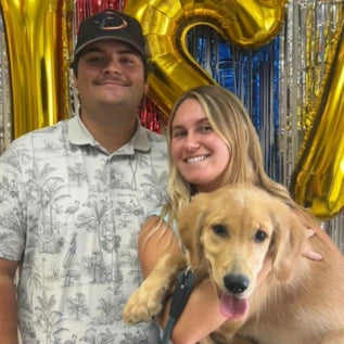 A smiling couple stands in front of foil streamers and large gold balloons with a Golden Retriever puppy. The man wears a patterned shirt and a cap, while the woman holds the puppy, which has its tongue out.