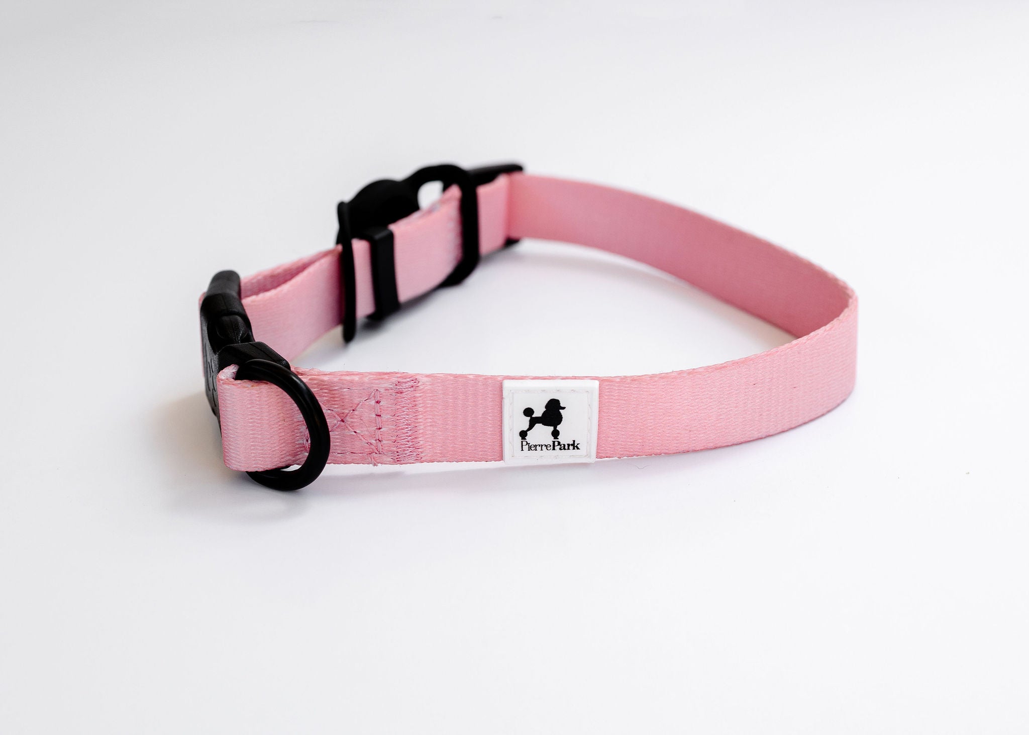 The Pink Dog Collar by PierrePark is displayed on a white surface, featuring a black plastic buckle, a metal D-ring, and a rectangular white label adorned with the silhouette of a dog.