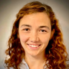 Smiling person with long, curly red hair against a neutral background. They are wearing a light-colored top.