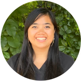 Smiling person with long dark hair, wearing earrings, in front of a leafy green background.