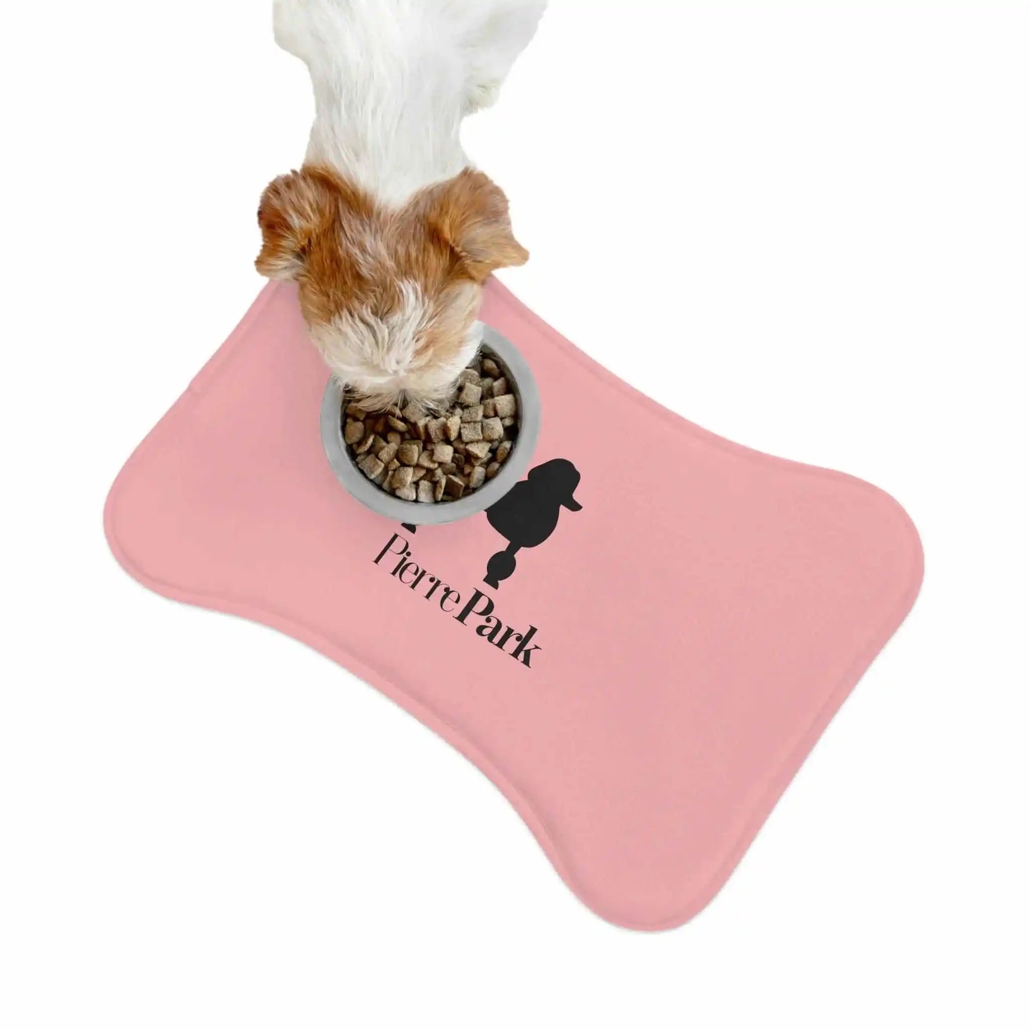 A small dog with white and brown fur eagerly eats kibble from a bowl. The Dog Food Mat - Pink by PierrePark features a dogs head silhouette and text Pierre Park, providing a non-slip bottom to keep everything in place.