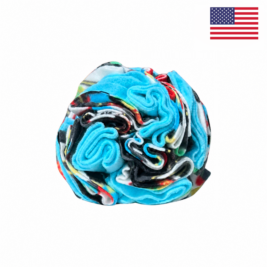A vibrant swirl of folded fabric in shades of blue, black, red, and white rests on a white background. The top right corner features a small American flag, emphasizing that the PierrePark Snuffle Ball for Dogs - Blue Games is proudly made in the USA.
