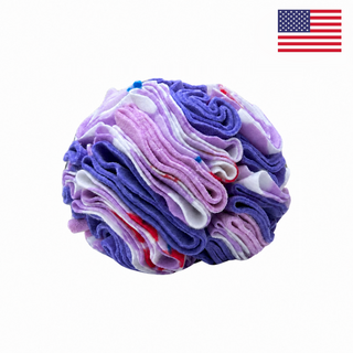 A Snuffle Ball for Dogs - Purple Cherries by PierrePark, featuring layered purple, pink, and white fabric strips, sits on a white background. Perfect for mental enrichment, this interactive puzzle toy promises hours of fun. A small US flag appears in the top right corner.